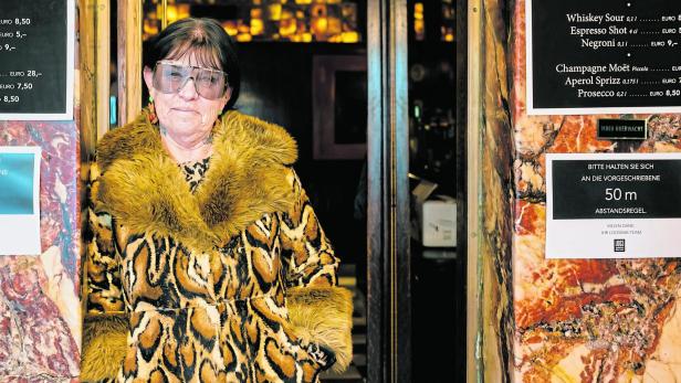 Eine ältere Frau mit großer Brille und auffälligem Mantel mit Fellkragen steht vor einem Eingang mit Marmorsäulen und Hinweisschildern.