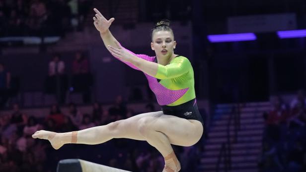 Eine Turnerin balanciert konzentriert auf einem Schwebebalken und führt eine Übung mit ausgestreckten Armen aus.