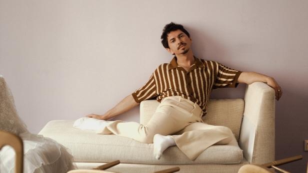 Ein Mann mit braun gestreiftem Hemd und heller Hose, Julian le Play, liegt entspannt auf einem cremefarbenen Sofa.