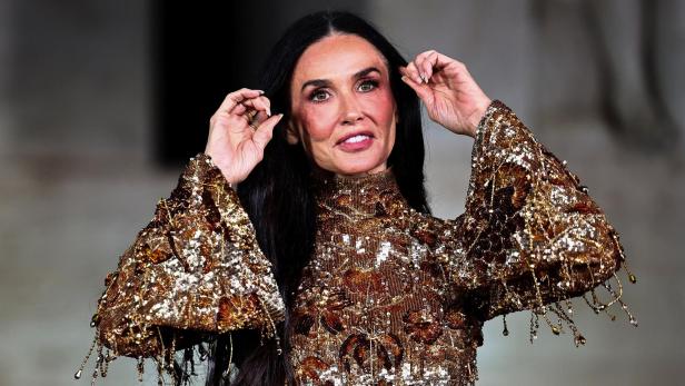 Demi Moore mit langen, dunklen Haaren trägt ein goldenes, paillettenbesetztes Oberteil und berührt ihr Haar.