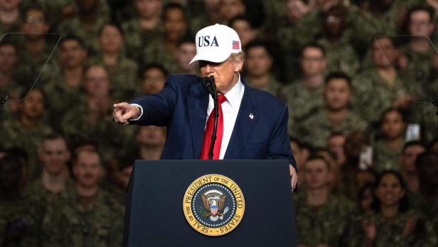 Donald Trump spricht vor einer Menschenmenge in Uniform, eine USA-Kappe tragend und auf etwas deutend.