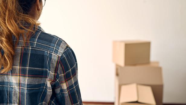 Eine Person mit Pferdeschwanz steht vor mehreren gestapelten Umzugskartons in einem leeren Raum.