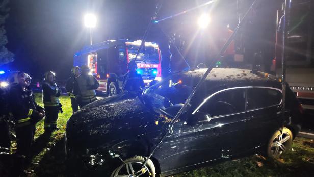 Mehrere Feuerwehrleute stehen nachts neben einem beschädigten Auto, das mit Seilen gesichert wird, während ein Einsatzfahrzeug leuchtet.