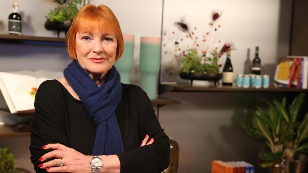 Chris Lohner Eine Frau mit rotem Haar und blauem Schal steht mit verschränkten Armen vor einem Regal mit Pflanzen, Büchern und Flaschen.