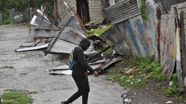 Eine Person mit Kapuze und Rucksack geht an zerstörten Wellblechzäunen und einem beschädigten Haus entlang einer nassen Straße vorbei.
