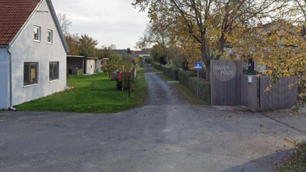 Eine schmale Straße führt zwischen einem weißen Haus und einem Holzzaun mit dem Schild „Bauer Pöltl“ in eine ländliche Gegend.