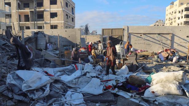 Mehrere Menschen durchsuchen Trümmer und Habseligkeiten vor zerstörten Gebäuden in einer städtischen Ruinenlandschaft.