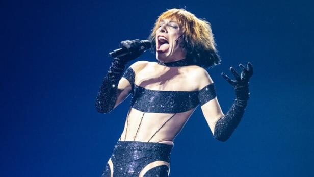 Nemo Nemo in glitzerndem Bühnenoutfit performt mit Mikrofon vor blauem Hintergrund.