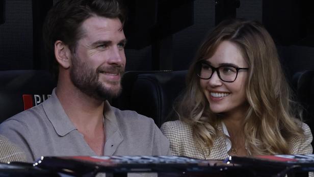 Liam Hemsworth Liam Hemsworth mit Bart und seine Partnerin Gabriella mit Brille sitzen lächelnd nebeneinander.