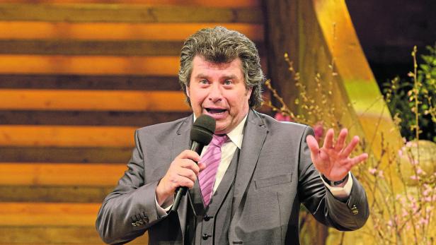 Ein Mann im Anzug spricht mit einem Mikrofon in der Hand und erhobener Hand vor einer Holztreppe.