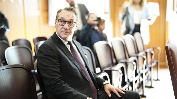 Strache im Anzug sitzt allein auf einer Stuhlreihe in einem hellen Raum, im Hintergrund sind weitere Personen unscharf zu sehen.