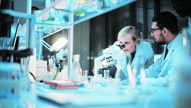 Medical Development Laboratory: Team of Female and Male Scientist Using Microscope, Analyzes Petri Dish Sample. Specialists Working on Medicine, Biotechnology Research in Advanced Pharma Lab Im Labor untersucht eine Wissenschaftlerin mit einem Mikroskop eine Probe, während ein Kollege zusieht.