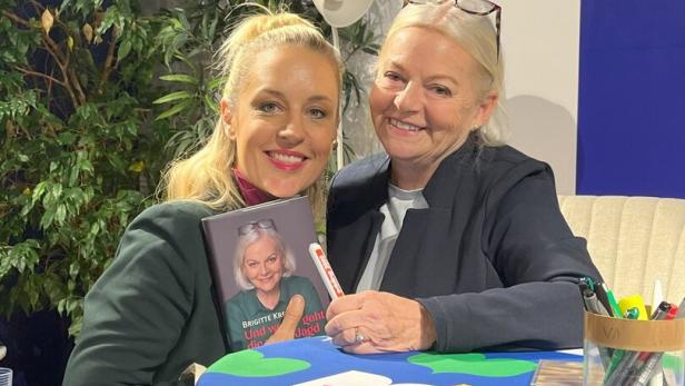 46-219829659 Zwei Frauen sitzen lächelnd zusammen, eine hält ein Buch mit dem Porträt der anderen, auf einem bunt gedeckten Tisch.