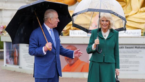 König Charles und Königin Camilla stehen im Regen unter Schirmen vor einem Denkmal.