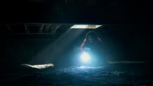 Eine Person kriecht mit einer Taschenlampe durch einen dunklen, staubigen Kellerraum, während Licht von oben einfällt.