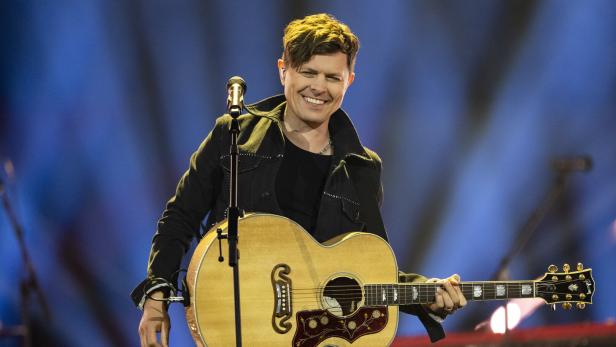 Michael Patrick Kelly spielt auf einer Bühne eine akustische Gitarre und steht vor einem Mikrofon.