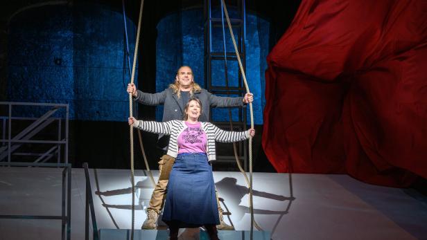 46-219864711 Zwei Personen stehen auf einer Bühne, halten Seile und lächeln, während im Hintergrund ein großes rotes Tuch hängt.