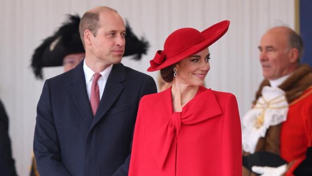 William und Kate Kate, in rotem Mantel und Hut, steht neben William bei einer Veranstaltung.