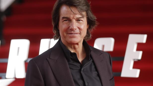 Tom Cruise  Tom Cruise posiert in einem dunklen Anzug vor einem roten Hintergrund mit weißen Buchstaben.