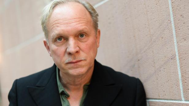 Schauspieler Ulrich Tukur Ulrich Tukur mit hellem Haar und Schnurrbart lehnt an einer Wand.