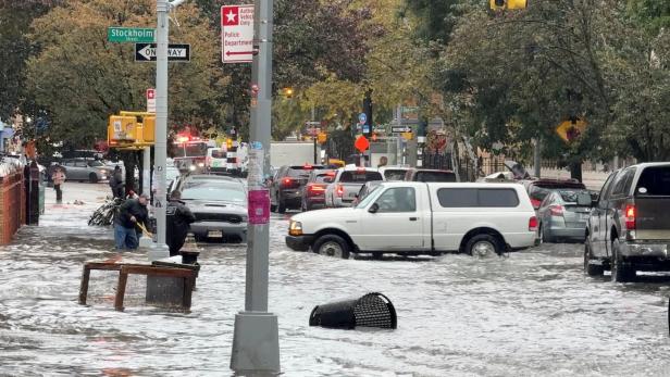 Vehicles move through floodwater in New York Straße mit überflutetem Wasser, Autos fahren langsam durch das Wasser, Menschen versuchen, den Gehweg zu reinigen.