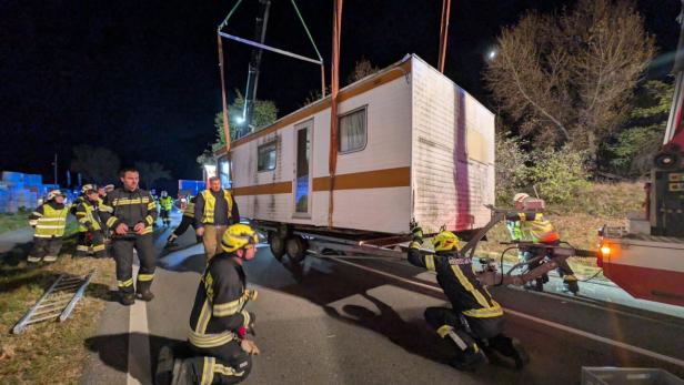 46-219882245 Mehrere Feuerwehrleute heben nachts mit einem Kran einen Wohnwagen von der Straße, während andere absichern.