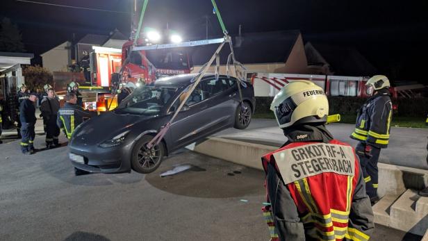 Ein Auto wird nachts von der Feuerwehr mit einem Kran angehoben, mehrere Feuerwehrleute beobachten den Einsatz.