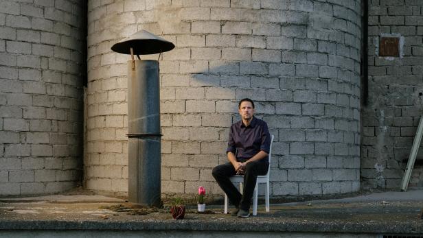 Ein Mann sitzt auf einem Stuhl vor einer grauen Backsteinwand, neben ihm stehen zwei Blumentöpfe.
