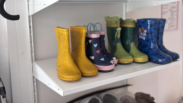 Mehrere Paar Gummistiefel und Sandalen stehen ordentlich auf Regalbrettern, darüber liegen zusammengefaltete Regenschirme.