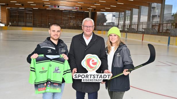 Drei Personen stehen auf einer Eishockeyfläche, halten ein Trikot, ein Schild mit „Sportstadt Eisenstadt“ und einen Eishockeyschläger.