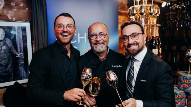 46-219884673 Drei Männer in festlicher Kleidung stoßen mit goldenen Sektgläsern an und lächeln in die Kamera.