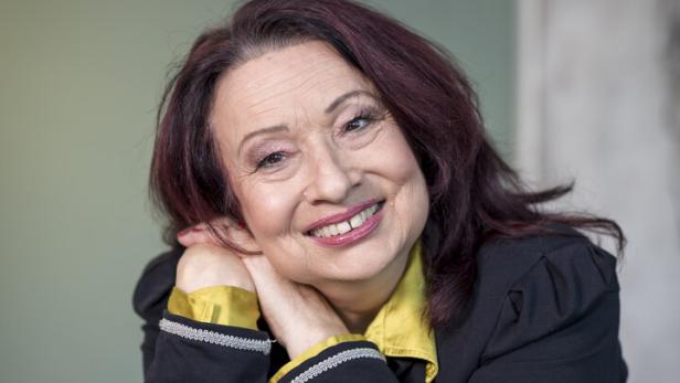 Sabine Holzinger Eine lächelnde Frau mit kastanienbraunen Haaren und gelber Bluse lehnt an einem Tisch.