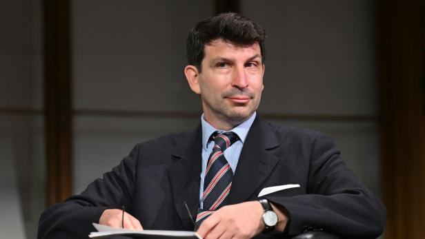 Ein Mann im Anzug sitzt auf einem Sessel, hält einen Block und schaut nachdenklich zur Seite.