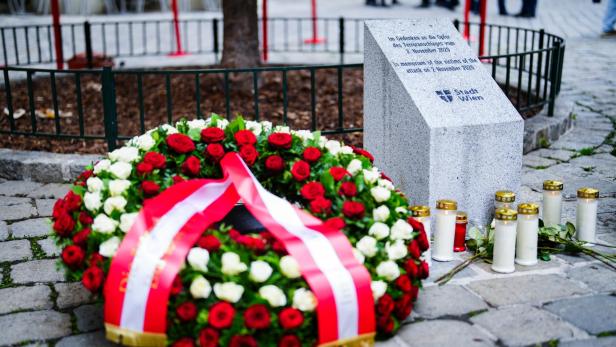 Ein Kranz mit rot-weißer Schleife liegt vor einem Denkmal der Stadt Wien, umgeben von Kerzen.