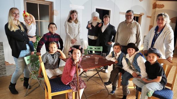 46-219781545 Mehrere Kinder und Erwachsene in verschiedenen Kostümen sitzen und stehen um einen Tisch in einem Raum mit Kulisse.