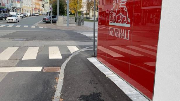 Ein schmaler Gehweg führt an einer roten Wand mit Generali-Logo entlang, daneben eine Kreuzung mit Ampeln und Zebrastreifen.