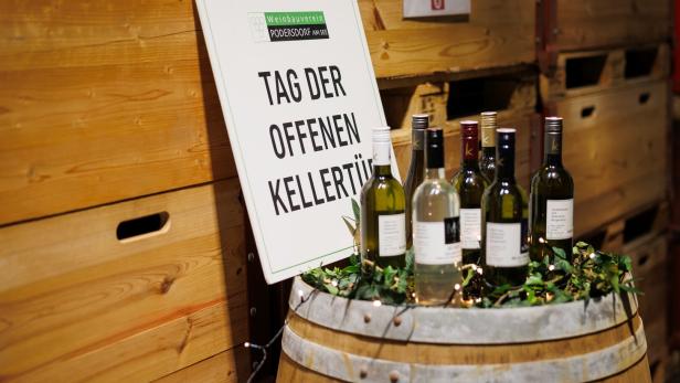 Mehrere Weinflaschen stehen auf einem Weinfass, daneben ein Schild mit der Aufschrift „Tag der offenen Kellertür“.