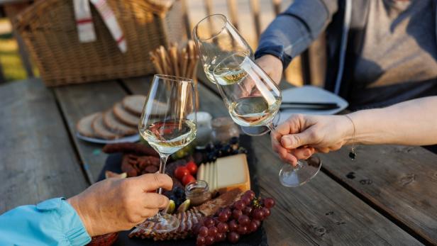 Drei Personen stoßen mit Weißweingläsern an, auf dem Tisch stehen Käse, Trauben, Brot und Wurst.