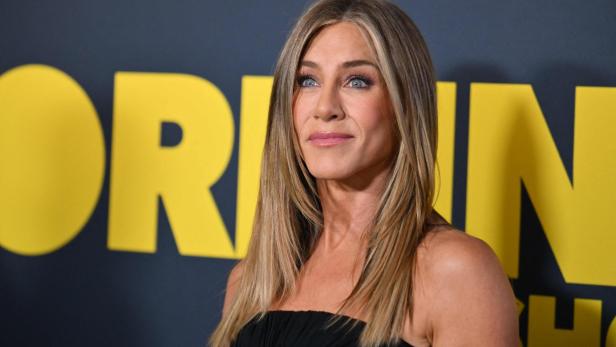 Jennifer Aniston posiert vor einem gelben Schriftzug auf dunklem Grund.