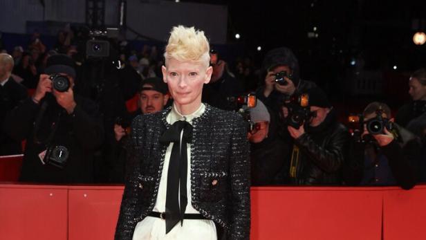 Eine Person mit kurzem, blondem Haar trägt eine gemusterte Hose, eine helle Bluse mit schwarzer Schleife und eine schwarze Jacke auf rotem Teppich.