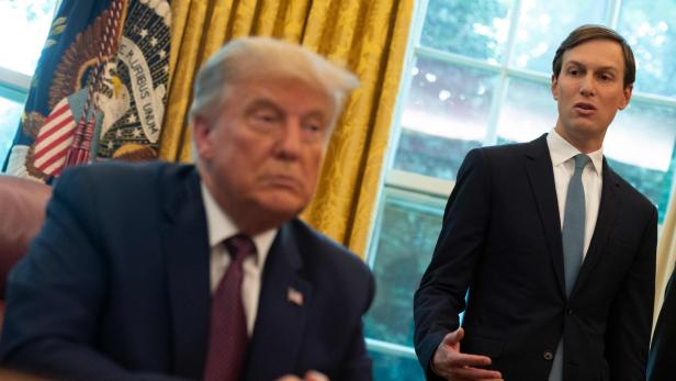 Donald Trump sitzt am Schreibtisch, während Jared Kushner im Anzug spricht.