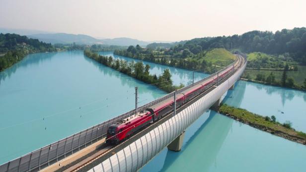 Ein roter Zug (Railjet) fährt über eine Brücke über einen türkisen Fluss 