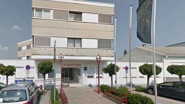 Ein modernes Rathaus mit flachem Dach, davor Bänke, Bäume, Fahnenmasten und geparkte Autos.