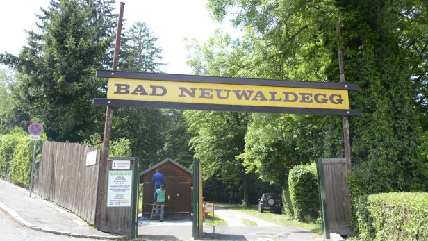 Ein Schild mit der Aufschrift "Bad Neuwaldegg" überspannt die Einfahrt, während zwei Personen an einem Häuschen arbeiten.