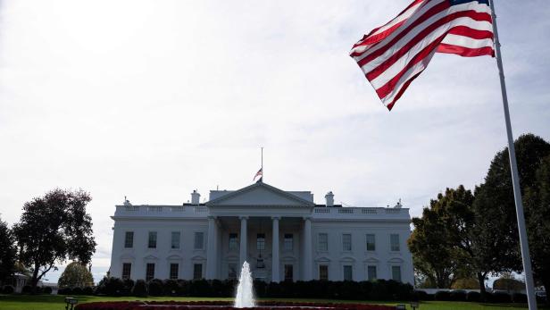 Das Weiße Haus mit amerikanischer Flagge im Wind und einem Springbrunnen davor.