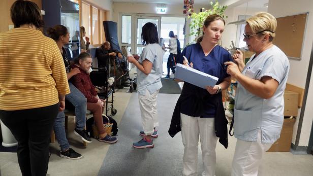 Mehrere Pflegekräfte und ältere Menschen befinden sich in einem Flur, einige sitzen, andere stehen und unterhalten sich.
