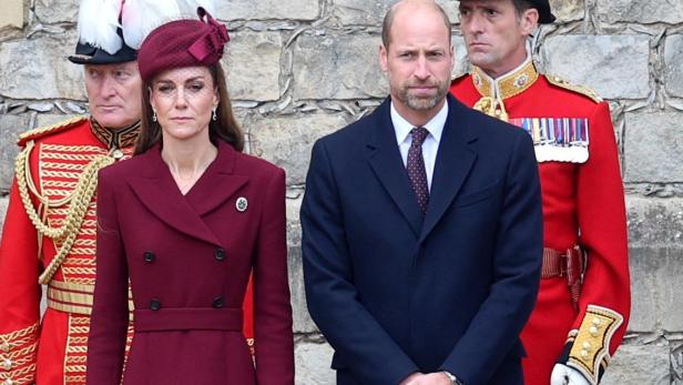 Kate und William stehen vor einer Steinmauer, flankiert von zwei Wachen in roten Uniformen.
