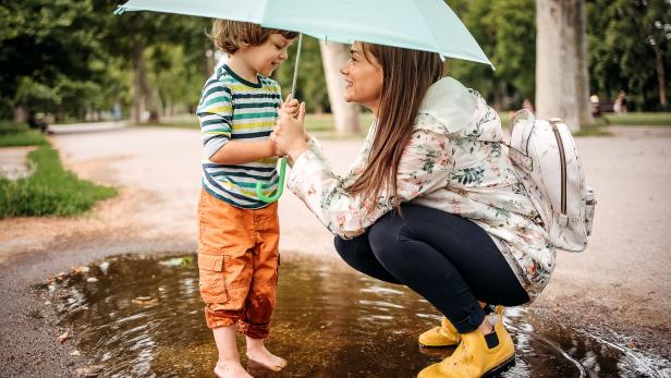 Eine Frau und ein Kind stehen gemeinsam unter einem Regenschirm und lachen, während das Kind barfuß in einer Pfütze steht.