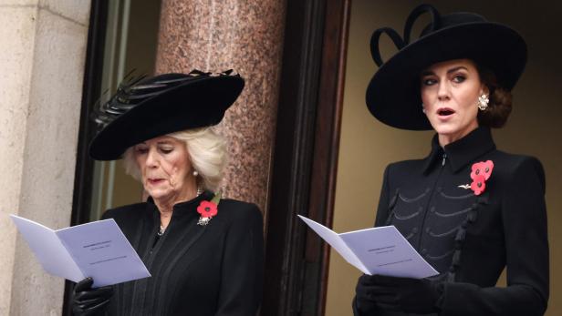 Camilla und Kate, beide mit schwarzen Hüten und Mohnblumen, lesen von Zetteln.