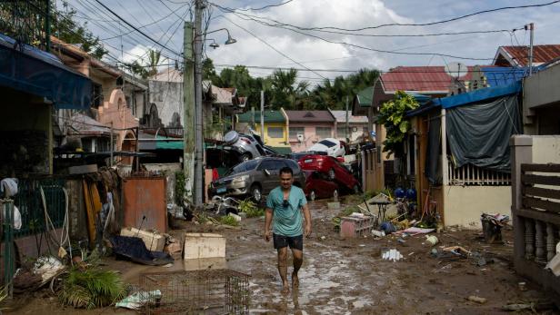 Ein Mann geht barfuß durch eine schlammige, von Überschwemmung und zerstörten Autos gezeichnete Straße mit Trümmern.